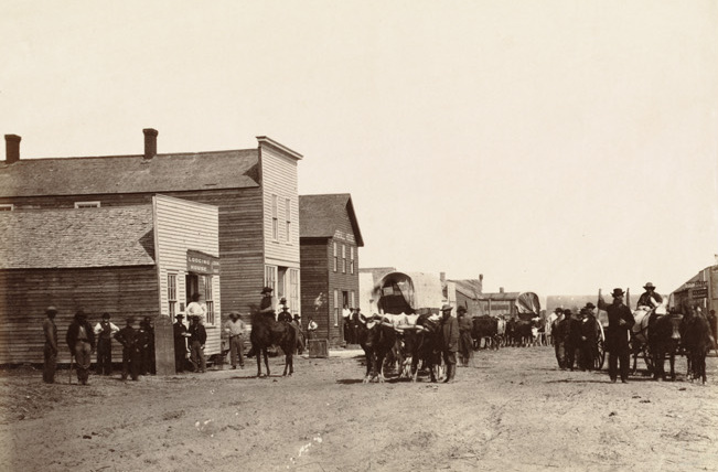 Wagons on a dirt path through a street with three buildings